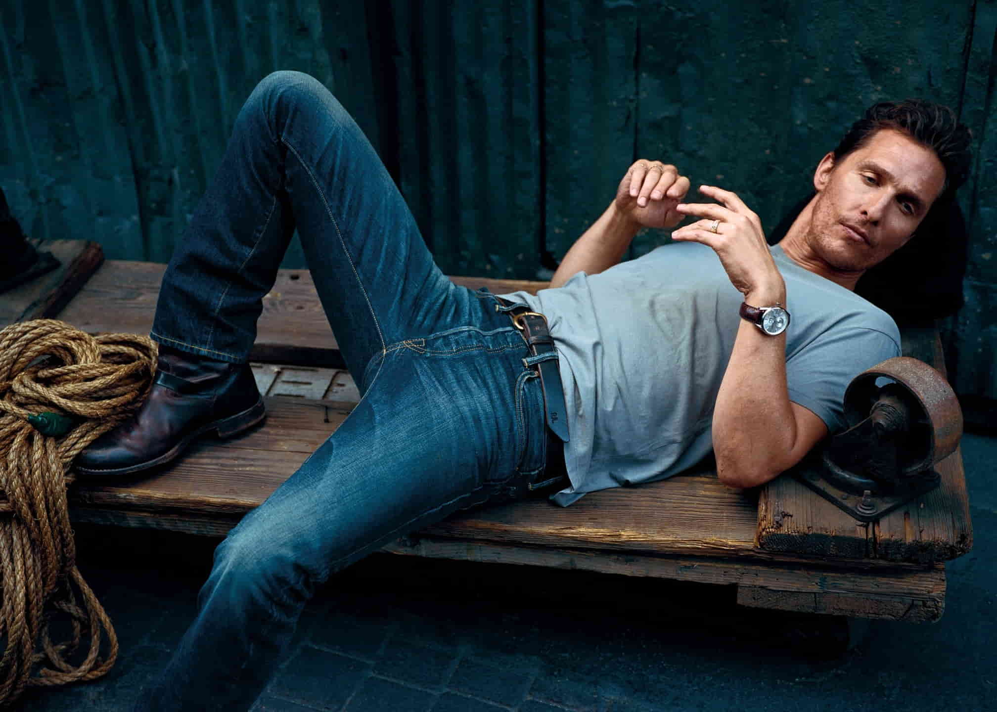 Matthew McConaughey in a blue t-shirt and denim jeans, posing by laying on a wooden platform