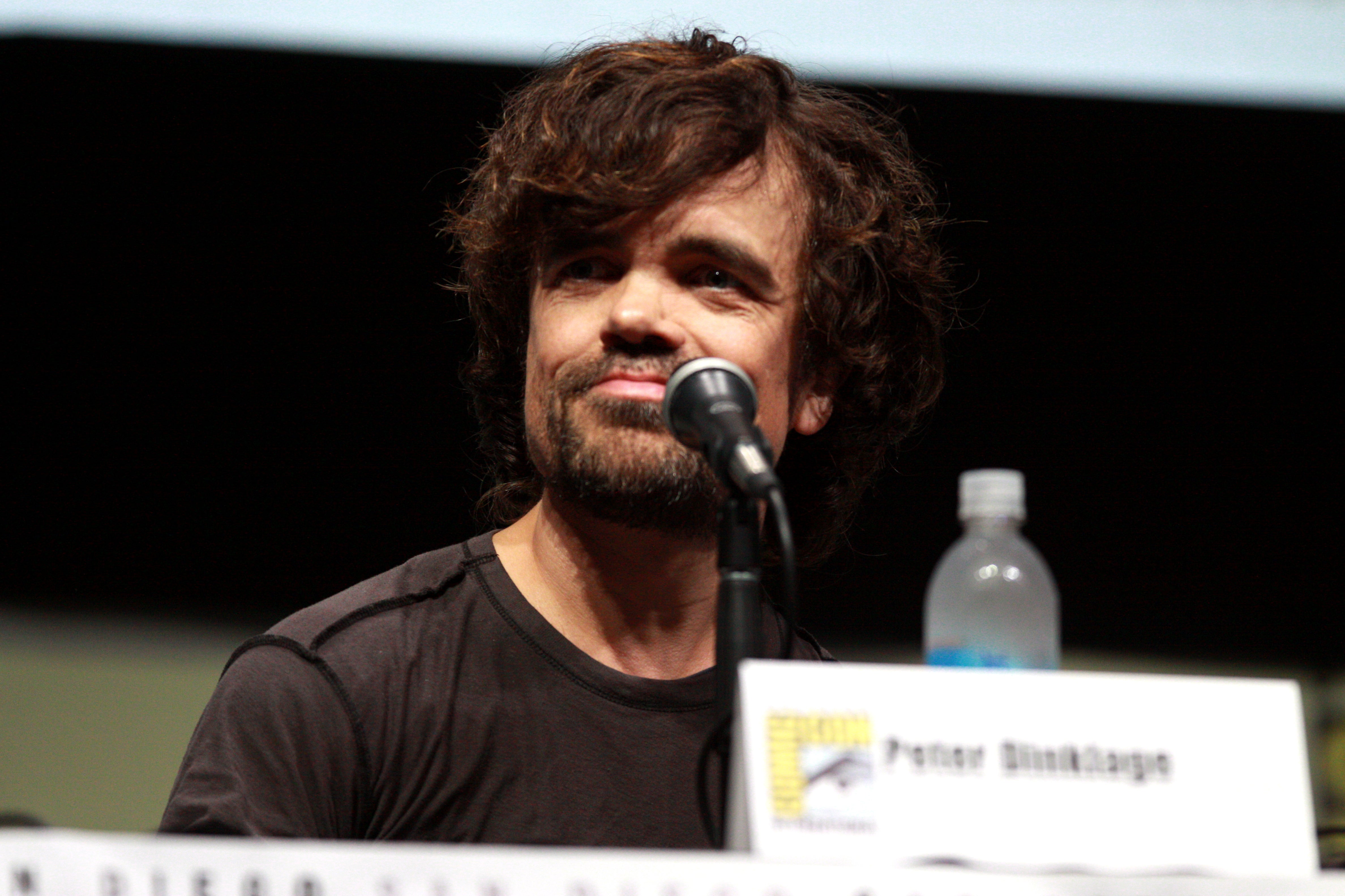 Peter Dinklage looking confident, with short hair and a dark t-shirt, addressing the audience at the Comicon event.