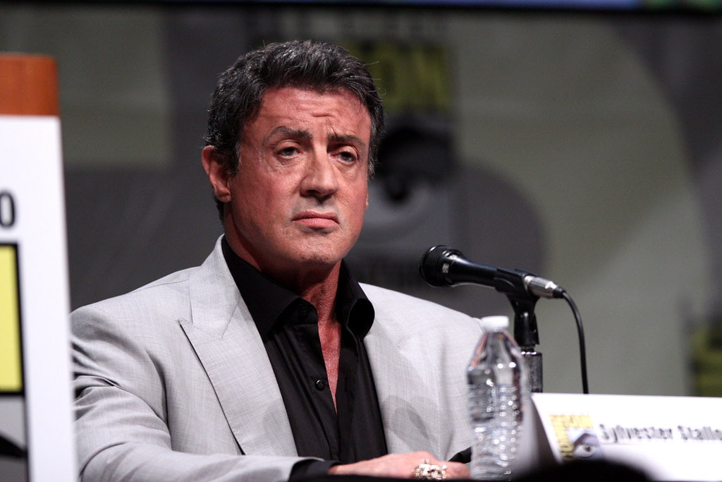 Sylvester Stallone wearing a grey suit and a black t-shirt setting at a press conference with a mic kept in front of him