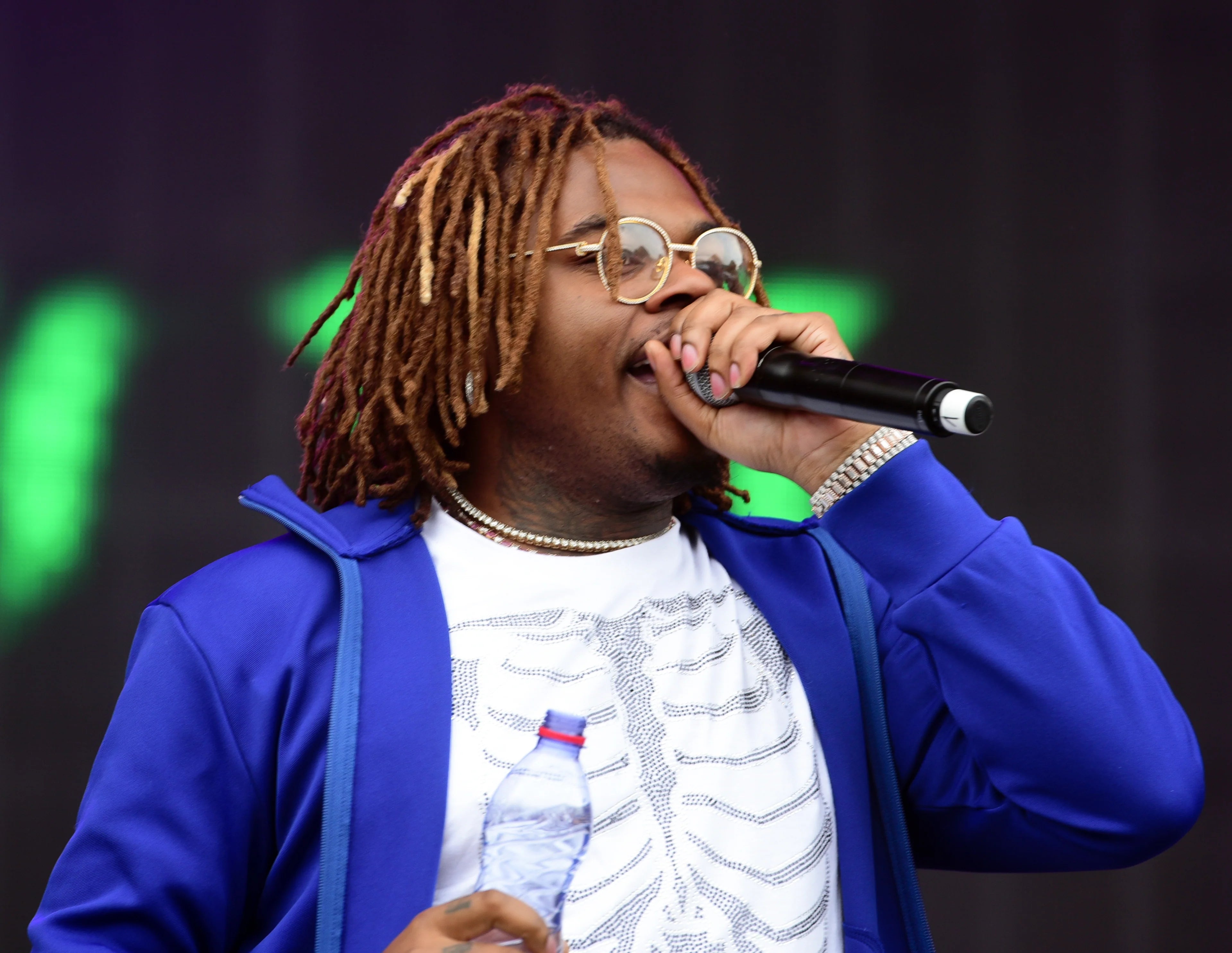 Gunna is wearing a white shirt and a blue jacket holding a microphone at a concert.